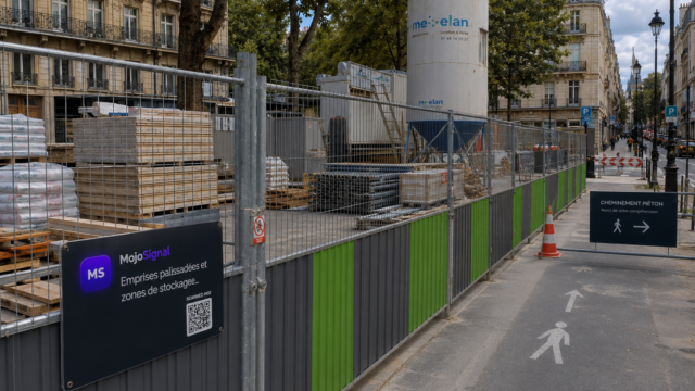 Emprise palissadée en ville avec stockage de matériaux de chantier et panneau de cheminement piéton.