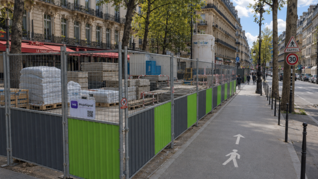 Longue zone de chantier palissadée sur trottoir parisien avec stockage de matériaux et passage piéton balisé.